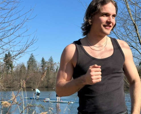 Die von Ihnen beschriebenen Bilder vermitteln eine heitere und fröhliche Atmosphäre. Die Person im schwarzen Tanktop mit ihrem strahlenden Lächeln vermittelt Glück und Zufriedenheit, während sie den Tag am See genießt. Die Paddleboarder auf dem Wasser verleihen der Szene ein aktives und erholsames Element und lassen vermuten, dass dies ein einladender Ort für Outdoor-Fans ist. Die kahlen Bäume deuten darauf hin, dass es früher Frühling oder später Herbst sein könnte, wenn die Bäume ihre Blätter abgeworfen haben, das Wetter aber noch angenehm genug für Outdoor-Aktivitäten ist. Der klare blaue Himmel verstärkt das allgemeine Gefühl der Ruhe und Gelassenheit in dieser malerischen Umgebung.