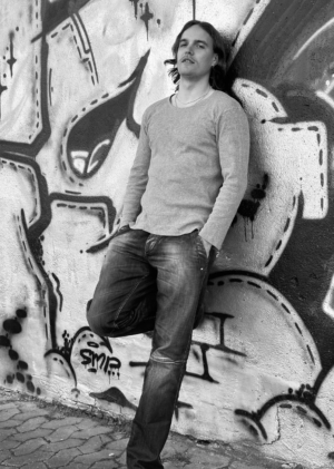 Dieses Schwarz-Weiß-Bild vermittelt urbane Coolness und Introspektion. Die Person in Langarmshirt und Jeans steht vor der Graffiti-bedeckten Wand, die als schlichte, künstlerische Kulisse dient. Die kräftigen Muster und abstrakten Designs des Graffitis verleihen der Szene Struktur und Kontrast und schaffen so eine faszinierende visuelle Erzählung. Der gepflasterte Boden verstärkt die raue, urbane Atmosphäre zusätzlich. Insgesamt vermittelt die Komposition einen Moment der Kontemplation inmitten einer dynamischen Stadtlandschaft, in der Kunst auf Alltag trifft.