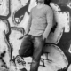 Dieses Schwarz-Weiß-Bild vermittelt urbane Coolness und Introspektion. Die Person in Langarmshirt und Jeans steht vor der Graffiti-bedeckten Wand, die als schlichte, künstlerische Kulisse dient. Die kräftigen Muster und abstrakten Designs des Graffitis verleihen der Szene Struktur und Kontrast und schaffen so eine faszinierende visuelle Erzählung. Der gepflasterte Boden verstärkt die raue, urbane Atmosphäre zusätzlich. Insgesamt vermittelt die Komposition einen Moment der Kontemplation inmitten einer dynamischen Stadtlandschaft, in der Kunst auf Alltag trifft.