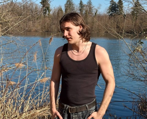 In dieser Szene strahlt die langhaarige Person am See eine besinnliche und gelassene Stimmung aus. Ihr schwarzes Tanktop und ihre Jeans ergänzen die Ruhe der Umgebung, verschmelzen subtil mit der Kargheit der kahlen Bäume und spiegeln ihre nachdenkliche Natur wider. Der klare blaue Himmel verstärkt diese friedliche Atmosphäre und wirft einen beruhigenden blauen Spiegel auf das ruhige Seewasser. Die Pose mit den Händen in den Taschen suggeriert einen Moment der Introspektion oder friedlichen Einsamkeit inmitten der schlichten Schönheit der Natur. Diese Bilder fangen eine perfekte Mischung aus menschlichen Emotionen und natürlicher Ruhe ein.