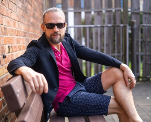 Das von Ihnen beschriebene Bild präsentiert eine faszinierende Gegenüberstellung von Modeelementen und Umgebungen. Maurice Leanders selbstbewusste Pose, zusammen mit seiner Wahl eines rosa Hemdes, kombiniert mit einem dunklen Blazer und gemusterten Shorts, zeugt von einem mutigen und vielseitigen Stilgefühl. Bart und Sonnenbrille unterstreichen sein lässiges und zugleich selbstbewusstes Auftreten. Die Backsteinmauern und der Metallzaun im Hintergrund verleihen der Szene einen urbanen Touch und ergänzen sein einzigartiges Outfit. Diese Kombination schafft eine visuell interessante Komposition, die Individualität und Selbstdarstellung unterstreicht.