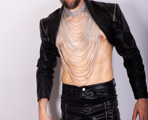 Die Beschreibung zeichnet das Bild einer Person mit einer mutigen, kantigen Ästhetik, die an den Stil von Maurice Leander erinnert. Die langen, zurückgebundenen Haare verleihen dem Look einen eleganten und selbstbewussten Touch und betonen die Gesichtszüge. Die schwarze Lederjacke und -hose vermitteln ein Gefühl von Härte und Rebellion, während die mehrlagigen Silberketten dem Outfit Struktur und Komplexität verleihen und optisch interessant wirken. Die Pose vor einem schlichten weißen Hintergrund lenkt die Aufmerksamkeit auf die Person und ihre auffälligen Modeentscheidungen und unterstreicht ihren einzigartigen Stil.