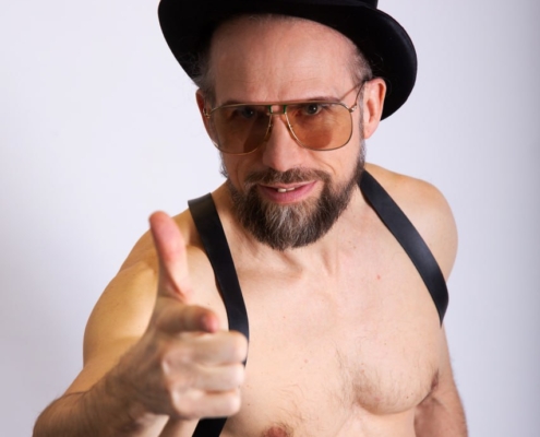 Die beschriebene Szene zeichnet ein eindrucksvolles und einprägsames Bild. Der bärtige Mann wirkt mit seiner einzigartigen Kombination an Accessoires mutig und kunstvoll. Sein hoher schwarzer Zylinder verleiht ihm einen Vintage- bzw. klassischen Stil, während die Sonnenbrille ihm einen modernen bzw. kantigen Touch verleiht. Oben ohne und mit schwarzen Hosenträgern verbindet er Formalität mit Lässigkeit und erzeugt so einen faszinierenden Kontrast, der Aufmerksamkeit erregt. Seine selbstbewusste Pose mit erhobenem Daumen signalisiert Selbstbewusstsein und positive Einstellung. Vor dem schlichten weißen Hintergrund betont die Gesamtkomposition seine markanten Gesichtszüge und seine Kleidung ohne Ablenkung. Das Bild zelebriert Individualität und persönlichen Ausdruck in seiner Schlichtheit und dennoch lebendigen Ausstrahlung.