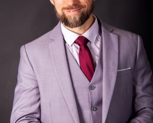 Das von Ihnen beschriebene Bild zeigt Maurice Leander als charmante und charismatische Persönlichkeit. Sein warmes Lächeln und seine selbstbewusste Haltung zeugen von einer einladenden und zugänglichen Persönlichkeit. Die Kombination aus hellgrauem Anzug und kastanienbrauner Krawatte präsentiert ihn professionell und dennoch stilvoll, wobei Bart und Pferdeschwanz ihm einen Hauch von Individualität verleihen. Der dunkle Hintergrund unterstreicht seine Präsenz und lässt ihn im Bild deutlicher hervortreten. Insgesamt vermittelt diese Darstellung sowohl Professionalität als auch persönlichen Stil.