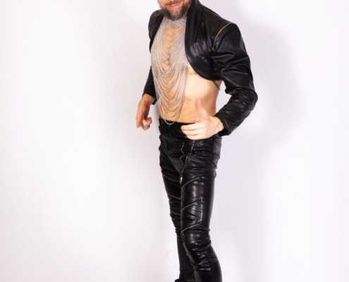 Maurice Leander verkörpert einen mutigen und kantigen Stil und strahlt durch seine selbstbewusste Pose mühelos Charisma aus. Der weiße Hintergrund unterstreicht seine Outfitwahl – eine markante Kombination aus Lederjacke und -hose, gepaart mit stylischen schwarzen Stiefeln. Seine nackte Brust wird von einer Halskette geschmückt, die ihm einen rebellischen Touch verleiht. Der Haarknoten rundet seinen Look ab und unterstreicht die insgesamt kantige Ästhetik, die er präsentiert. Dieses eindrucksvolle Bild fängt Maurices einzigartigen Sinn für Mode und Persönlichkeit ein.