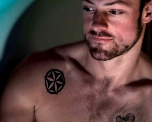 Die von Ihnen beschriebenen Bilder vermitteln ein Gefühl von Introspektion und Charakter. Die Kombination der körperlichen Merkmale des Mannes, wie sein Bart und die Tattoos – ein Kompassstern und ein Anker – suggerieren ein Thema der Navigation und Erdung. Diese Symbole werden oft mit Orientierung, Erkundung, Stabilität und Stärke assoziiert. Das gedämpfte Licht verstärkt diese Stimmung, indem es bunte Schatten wirft, die über seine Gestalt spielen und der kontemplativen Atmosphäre Tiefe verleihen. Diese Umgebung hebt nicht nur seine körperlichen Merkmale hervor, sondern deutet vielleicht auch auf eine innere Reise oder Reflexion hin. Sollte diese Beschreibung Teil eines Kunstwerks oder Fotos sein, das Sie in Betracht ziehen, fängt sie sowohl das raue Äußere ein, das üblicherweise mit einer „Callboy“-Persönlichkeit assoziiert wird, als auch Elemente der Nachdenklichkeit und Verletzlichkeit durch Ausdruck und Licht ein.