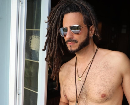 Die von Ihnen beschriebene Szene vermittelt ein lebendiges Bild einer entspannten, besinnlichen Atmosphäre. Der Mann mit seinen markanten langen Dreadlocks und der Sonnenbrille strahlt eine coole und entspannte Ausstrahlung aus. Sein oberkörperfreies Auftreten suggeriert Geborgenheit und Gelassenheit, vielleicht einen Moment der Selbstbesinnung oder einfach nur den Augenblick genießend. Die Halskette verleiht ihm einen persönlichen Stil, während die kleine Tasse, die er hält, darauf hindeuten könnte, dass er einen Drink genießt oder einem zwanglosen Ritual nachgeht. Das Stehen neben einer offenen Tür könnte Offenheit für neue Möglichkeiten oder den Übergang zwischen verschiedenen Räumen oder Gemütszuständen symbolisieren. Insgesamt ist es eine Szene, die sowohl Individualität als auch introspektive Ruhe einfängt.
