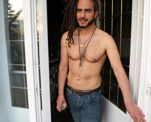Die Szene fängt eine entspannte und lockere Atmosphäre ein und spiegelt perfekt den unbeschwerten Geist wider, der üblicherweise mit Figuren wie Callboy Aslan assoziiert wird. Der oberkörperfreie Mann mit seinen langen Dreadlocks und dem Bart strahlt Freiheit und Lässigkeit aus. Seine Kleidung – schlichte Bluejeans – und die verschiedenen Halsketten um seinen Hals unterstreichen diese lockere Persönlichkeit zusätzlich. Die Getränkedose in seiner Hand suggeriert einen Moment der Muße oder des ungezwungenen Genusses, während seine Pose im Türrahmen Offenheit und Geborgenheit in diesem sonnendurchfluteten Raum vermittelt. Der geflieste Boden unter den Füßen trägt zu dem Gefühl von Wärme und Entspannung bei, das den Raum durchdringt. Insgesamt ist es eine Szene, die einen unbeschwerten Lebensstil verkörpert, der von Einfachheit und der Wertschätzung der kleinen Freuden des Lebens geprägt ist.