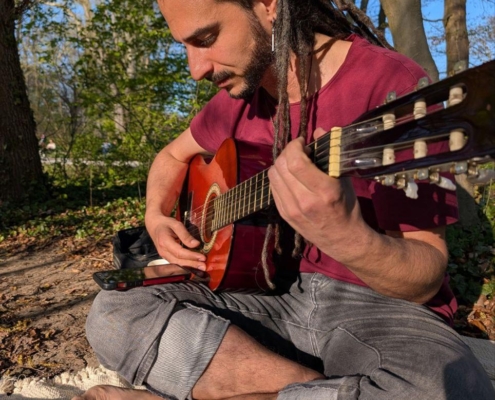 Die Szene fängt einen ruhigen und harmonischen Moment ein, in dem Callboy Aslan mit seinen langen, herabhängenden Dreadlocks in der natürlichen Schönheit des Parks Ruhe findet. Die Melodien der Akustikgitarre verschmelzen nahtlos mit dem sanften Rascheln der Blätter und den sanften Sonnenstrahlen, die durch das Blätterdach fallen. In einem kastanienbraunen Hemd und hochgekrempelten Jeans verkörpern sie eine entspannte Stimmung und tauchen in die wohltuende Umgebung ein. Diese ruhige Umgebung bietet die ideale Kulisse für kreativen Ausdruck und Reflexion, sodass Musik und Natur mühelos in dieses idyllische Tableau verschmelzen.