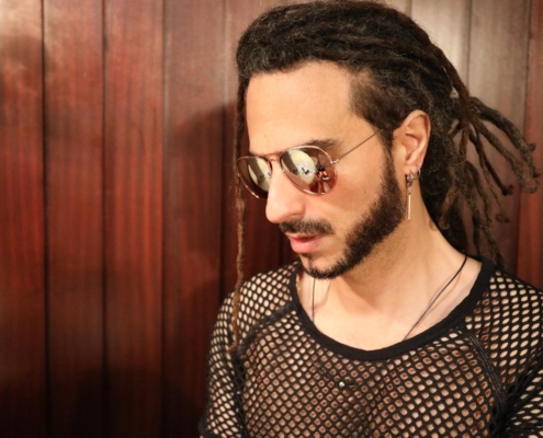 Die Beschreibung zeichnet das eindrucksvolle Bild einer Person mit markanten Gesichtszügen und geheimnisvoller Aura. Die Kombination aus Dreadlocks und Sonnenbrille sorgt für ein faszinierendes Element, während das schwarze Netzhemd auf eine mutige Modewahl schließen lässt. Die Spiegelung in der Sonnenbrille bietet Raum für künstlerische Interpretation und unterstreicht die Tiefe und den rätselhaften Charakter der Figur. Der hölzerne Hintergrund bildet einen natürlichen Kontrast zu ihrer modernen Kleidung und unterstreicht ihr nachdenkliches Auftreten. Dieses Bild könnte Neugier auf Callboy Aslans Geschichte und Persönlichkeit wecken.