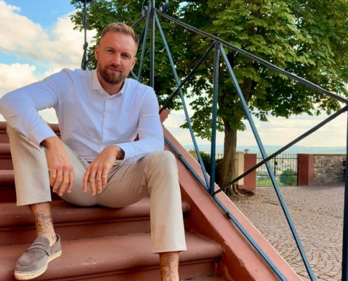 Ein Mann mit Tattoos, gekleidet in ein weißes Hemd und eine beigefarbene Hose, sitzt lässig auf einer Freitreppe. Hinter ihm bestimmen üppige Bäume und eine Straßenlaterne die Szene. Er wirkt entspannt, während er unter dem bedeckten Himmel nachdenklich über einen gepflasterten Hof auf einen entfernten Zaun blickt - eine nachdenkliche und ruhige Haltung im gedämpften Licht. Dies ist Callboy David, der in einem Moment der stillen Besinnung im Freien aufgenommen wurde.