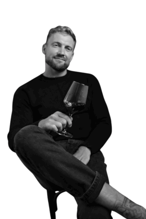Callboy David, ein Mann mit kurzen Haaren und Bart, sitzt in dunklem Pullover und Jeans auf einem Hocker, lächelt und hält einen Rotwein in der Hand.