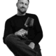 Callboy David, ein Mann mit kurzen Haaren und Bart, sitzt in dunklem Pullover und Jeans auf einem Hocker, lächelt und hält einen Rotwein in der Hand.