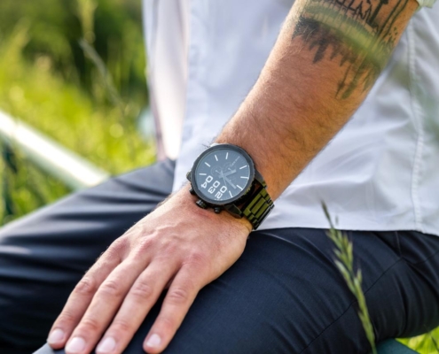 Callboy Michael lehnt sich lässig auf einem grünen Geländer im Freien zurück, gekleidet in ein frisches weißes Hemd und eine dunkle Hose. Seine Armbanduhr glänzt dezent, während er seinen Arm ablegt, und lässt das kunstvolle Tattoo der Skyline auf seinem Unterarm sichtbar werden – eine Mischung aus urbaner Kunstfertigkeit und persönlichem Stil. Hinter ihm verschwimmt eine üppige Landschaft aus grünem Gras und Bäumen sanft mit dem Hintergrund und ruft eine entspannte Sommeratmosphäre inmitten der Natur hervor, während Michaels gelassene Erscheinung urbane Eleganz erahnen lässt.