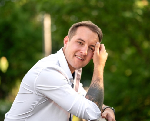 Michael, ein Callboy, steht draußen vor einem sanft verschwommenen grünen Hintergrund. Er trägt ein frisches weißes Hemd mit teilweise hochgekrempelten Ärmeln, die seine tätowierten Unterarme freigeben. Ein entspanntes Lächeln erhellt sein Gesicht, während er sich lässig mit der Hand durch die Haare fährt. Das Grün hinter ihm bildet einen ruhigen und frischen Kontrast zu seiner eleganten Kleidung und seiner tätowierten Haut und vermittelt ein lässiges und dennoch selbstbewusstes Auftreten.