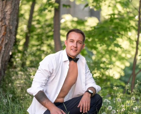 Callboy Michael kniet im üppigen Gras eines sonnendurchfluteten Waldes, durch dessen Bäume Sonnenlicht fällt. Sein weißes Hemd ist offen und gibt den Blick auf seine Brust und ein Tattoo frei, das sich entlang seines linken Unterarms schlängelt. Eine schwarze Fliege hängt locker um seinen Hals und harmoniert perfekt mit seiner gut sitzenden schwarzen Hose und der eleganten Uhr am Handgelenk. Mit einem leichten, selbstbewussten Lächeln blickt er direkt in die Kamera – sein Auftreten wirkt entspannt, aber einladend, während er inmitten der ruhigen Schönheit der Natur posiert.