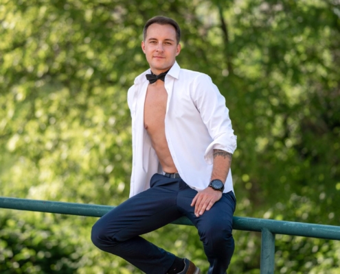 Michael, der Callboy, sitzt lässig auf einem grünen Metallgeländer im Freien, umgeben von gesprenkeltem Sonnenlicht, das durch üppige grüne Bäume fällt. Er trägt ein aufgeknöpftes weißes Hemd, das seine Brust freigibt, dazu elegante dunkle Hosen und polierte schwarze Schuhe. Eine ordentlich gebundene Fliege ziert seinen Hals und verleiht seiner entspannten Pose einen verspielten Hauch von Formalität. Sein Auftreten ist selbstsicher und doch zugänglich, während er in die Ferne oder auf den Betrachter blickt, umgeben von leuchtendem Grün und einer Atmosphäre sommerlicher Gelassenheit.
