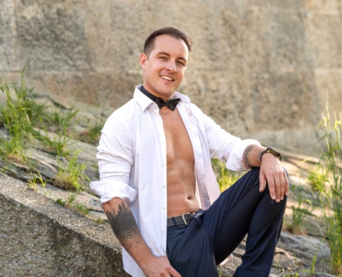 Sicher! Hier ist ein beschreibender Absatz basierend auf Ihren Angaben: Callboy Michael sitzt gemütlich auf einer abschüssigen Steinfläche im Freien, umgeben von hohem Gras, das zwischen den verwitterten Steinen sprießt. Er trägt dunkle Hosen und passende Schuhe, sein weißes Hemd ist halb aufgeknöpft und gibt den Blick auf seine tätowierte Brust frei. Eine schwarze Fliege hängt locker um seinen Hals. Trotz der rauen Umgebung verleihen Michaels unbeschwertes Lächeln und sein selbstbewusstes Auftreten der Szene Wärme, während das Sonnenlicht auf seiner tätowierten Haut und seinem frischen Outfit tanzt.