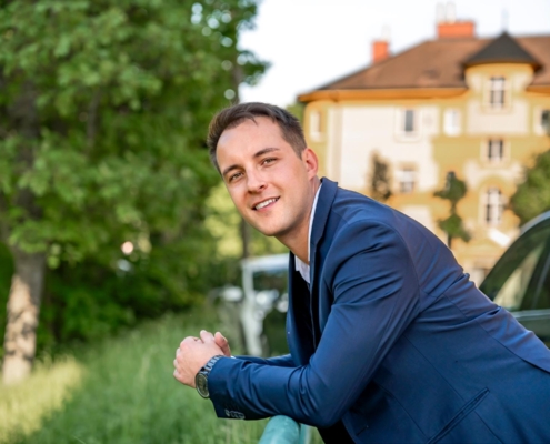 Natürlich! Hier ist eine anschauliche Zusammenfassung Ihrer Szene: Callboy Michael steht selbstbewusst da, gekleidet in einen schicken blauen Anzug, der einen schönen Kontrast zur Umgebung bildet. Lässig lehnt er an einem grünen Geländer, sein Gesichtsausdruck ist warm, während er direkt in die Kamera lächelt. Hinter Michael wiegen sich üppige Bäume sanft, deren Blätter das Sonnenlicht einfangen. Der Hintergrund ist leicht unscharf, lässt aber die hellbraunen und gelben Wände eines Gebäudes erkennen, die im hellen Tageslicht leuchten und die fröhliche und einladende Atmosphäre der Szene verstärken.