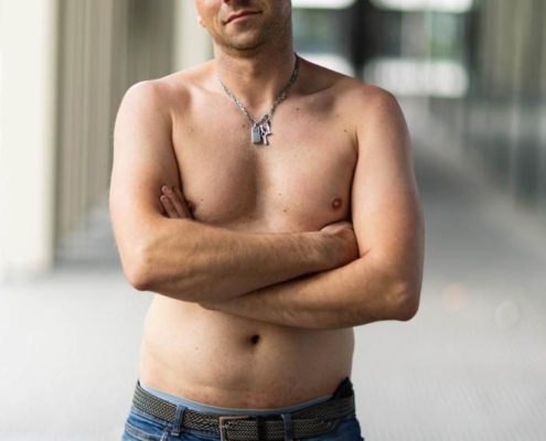 Auf jeden Fall! Hier ist eine anschauliche Zusammenfassung basierend auf Ihrer Frage: Callboy Matteo steht selbstbewusst in einem Flur. Sein kahler Kopf fängt das Umgebungslicht ein. Ein leichtes, wissendes Lächeln umspielt seine Lippen, während er ohne Hemd posiert und seine schlanke Figur betont. Seine Arme sind vor der Brust verschränkt, was sein selbstbewusstes Auftreten unterstreicht. Er trägt schlichte Jeans und eine dezente Halskette, die auf seiner Haut liegt. Im Hintergrund sind unscharfe Säulen zu sehen, die Matteo einrahmen und der Szene eine elegante Tiefe verleihen.