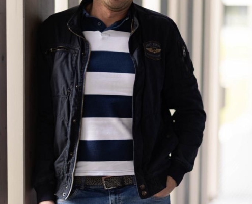 Ein Mann mit kurzen Haaren und gepflegtem Bart, der Callboy Matteo ähnelt, steht in einem modernen, gut beleuchteten Flur. Er trägt eine elegante schwarze Jacke über einem blau-weiß gestreiften Poloshirt und lässige Bluejeans. Sein entspanntes Lächeln strahlt Wärme aus, während er gemütlich an der Wand lehnt, beleuchtet von moderner Beleuchtung, die die klaren Linien und die Atmosphäre des Flurs unterstreicht.