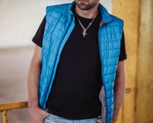 Callboy Matteo steht drinnen neben einem gelben Geländer und verkörpert eine nachdenkliche Präsenz. Sein rasierter Kopf und sein gepflegter Bart werden von der sanften Innenbeleuchtung erhellt. Er trägt eine blaue Pufferweste über einem schwarzen Hemd, kombiniert mit hellen Jeans, die seinen lässigen und doch unverwechselbaren Look vervollständigen. Mit seinem in die Ferne gerichteten Blick deutet Matteos Haltung auf stille Kontemplation hin, während er leicht am Geländer lehnt und die Atmosphäre seiner Umgebung in sich aufnimmt.