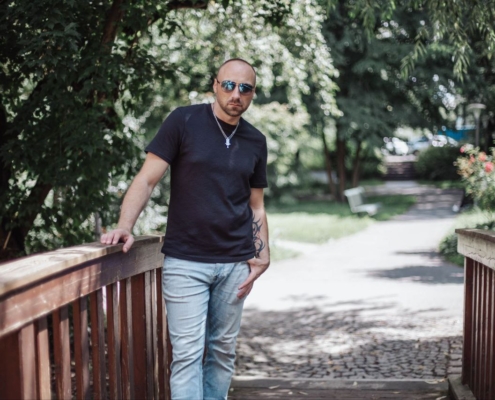 Matteo, ein Callboy mit selbstbewusstem Charme, steht lässig in der Mitte einer hölzernen Fußgängerbrücke, die in warmes Sonnenlicht getaucht ist. Er trägt eine stylische Sonnenbrille, die den blauen Himmel reflektiert, ein enges dunkles T-Shirt, das seine Figur betont, und hell gewaschene Jeans. Die Brücke unter seinen Füßen wölbt sich sanft über einen seichten Bach oder Pfad. Hinter ihm erstreckt sich ein einladender Park: Leuchtend grüne Bäume werfen gesprenkelte Schatten auf die Grasflächen und blumenübersäten Lichtungen. Sonnenlicht fällt durch die Blätter, beleuchtet Matteos Figur und schafft eine Atmosphäre entspannter Sommerlichkeit. Seine Haltung vermittelt sowohl Zugänglichkeit als auch stille Selbstsicherheit – perfekt zu Hause inmitten der üppigen Schönheit der Natur.