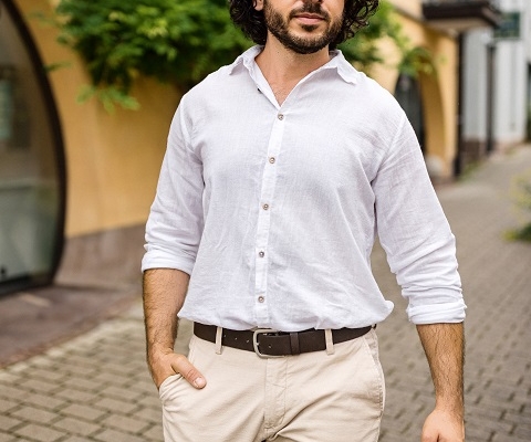 Ein Mann mit lockigem, dunklem Haar und Bart, der Callboy Kaan ähnelt, geht draußen auf einem gepflasterten Weg spazieren. Er trägt ein weißes Hemd und eine beige Hose. Auf der einen Seite des Weges steht ein gelbes Gebäude, und ringsherum ist viel Grün – Büsche oder vielleicht auch Kletterpflanzen –, was die sommerliche, entspannte Atmosphäre verstärkt. Der Gesamteindruck ist lässig und doch stilvoll, wobei das natürliche Licht die Szenerie unterstreicht.