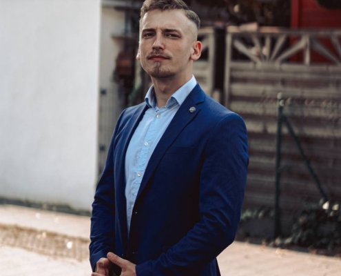 Ein Mann steht im Freien, gekleidet in einen blauen Anzug und ein hellblaues Hemd. Er hat die Hände vor dem Körper gefaltet und blickt selbstbewusst in die Kamera. Die Szene spielt im Freien, mit einem Holzzaun an der Seite, einem Tor und einem Gebäude im Hintergrund. Das natürliche Licht hebt seine Gesichtszüge hervor und verleiht seiner Haltung und seinem Ausdruck eine selbstbewusste Präsenz.