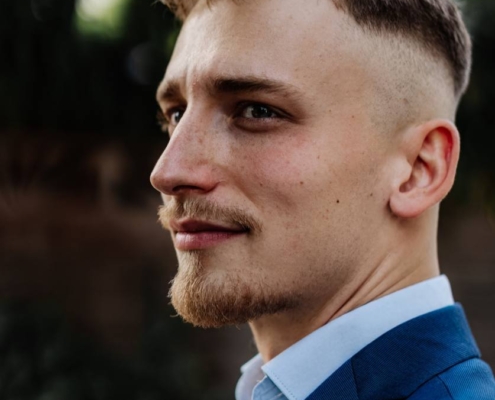 Ein junger Mann mit kurzen blonden Haaren und gepflegtem Bart, bekannt als Callboy Gabriel L., ist im Freien abgebildet. Er trägt einen eleganten blauen Anzug mit hellblauem Hemd, ohne Krawatte. Er blickt nachdenklich zur Seite, sein Gesichtsausdruck ist ruhig und selbstbewusst. Der Hintergrund ist mit weichgezeichnetem Grün gestaltet, das eine einladende und natürliche Atmosphäre schafft, die seinen gepflegten Look perfekt unterstreicht.