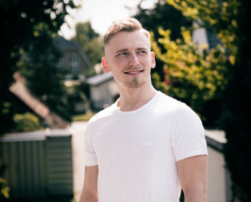 Ein junger Mann mit kurzen blonden Haaren und Bart steht im Freien, lässig gekleidet in weißem T-Shirt und Bluejeans. Er ist Callboy Gabriel L. und blickt lächelnd zur Seite. Sein Gesichtsausdruck ist entspannt und freundlich. Der Hintergrund ist sonnendurchflutet, Bäume und Stadtgebäude sind leicht unscharf und vermitteln den Eindruck eines angenehmen Tages in einem Stadtpark oder auf der Straße.