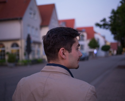 Es klingt, als würden Sie einen ruhigen, filmischen Moment beschreiben. Hier ist eine mögliche Erweiterung oder Interpretation, je nach Ihrer Absicht: Ein Mann mit kurzen braunen Haaren trägt eine hellbraune Jacke, als er in der Abenddämmerung eine ruhige Wohnstraße entlangschlendert. Die Häuser stehen ruhig zu beiden Seiten, ihre Fenster leuchten sanft im schwindenden Licht, während Bäume die Bürgersteige säumen und sanfte Schatten auf das Pflaster werfen. Der Himmel ist in düstere Rosa- und Blautöne getaucht. Der Mann blickt leicht nach links - vielleicht ist er in Gedanken versunken oder hat etwas bemerkt - eine friedliche Ruhe, die ihn und die ruhige Nachbarschaft um ihn herum einhüllt. Wenn Sie daraus etwas Bestimmtes machen möchten (z. B. einen Anfang für eine Geschichte, eine Fotobeschreibung oder eine Anregung für Kunst/AI-Bilderzeugung), lassen Sie es mich wissen!.