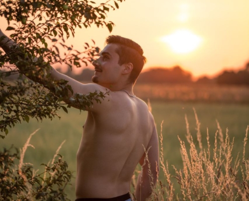 Ein Mann ohne Hemd steht in hüfthohem, goldenem Gras neben einem einsamen Baum, während die Sonne hinter ihm untergeht. Er ist leicht von der Kamera abgewandt, blickt aber mit einem warmen Lächeln über seine Schulter zurück. Die untergehende Sonne taucht die Landschaft in satte, goldene Farbtöne, wirft lange Schatten und erzeugt ein weiches Gegenlicht, das die Konturen seines Körpers hervorhebt und das hohe Gras um ihn herum beleuchtet. Der Himmel leuchtet in orangefarbenen und rosafarbenen Tönen und unterstreicht die heitere und fröhliche Stimmung dieser Dämmerungsszene in der Natur.