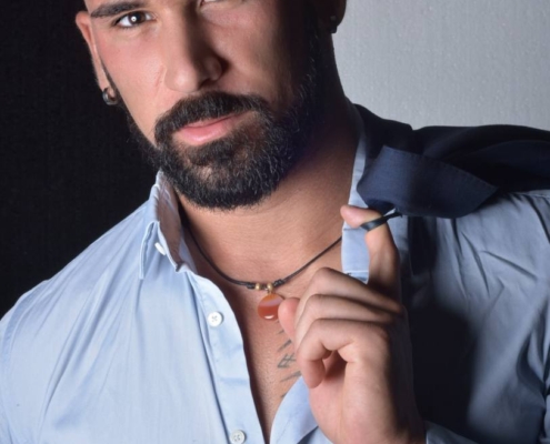 Ein Mann mit dunklem Haar und gepflegtem Bart steht selbstbewusst vor einem neutralen Hintergrund. Er trägt ein hellblaues Button-up-Hemd, dessen Ärmel lässig hochgekrempelt sind, und hält mit einer Hand ein Jackett über die Schulter gelegt. Sein Ausdruck ist selbstbewusst und strahlt einen mühelos stilvollen Charme aus, der an Callboy Amador erinnert. Der Gesamteindruck vermittelt Gelassenheit und moderne Raffinesse.