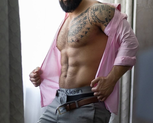 Ein bärtiger Mann mit Tätowierungen auf den Schultern steht selbstbewusst in einem Haus. Er lächelt, seine Augen leuchten und sind einnehmend, während er ein aufgeknöpftes rosa Hemd öffnet und eine straffe, muskulöse Brust zum Vorschein bringt. Der Bund seiner Calvin-Klein-Unterwäsche lugt über seine Hose hervor. Hinter ihm dringt sanftes Tageslicht durch die durchsichtigen Vorhänge und taucht die Szene in ein warmes Licht. Die Gesamtstimmung strahlt Kühnheit und Selbstbewusstsein aus, die an den Stil von Callboy Amador erinnern.
