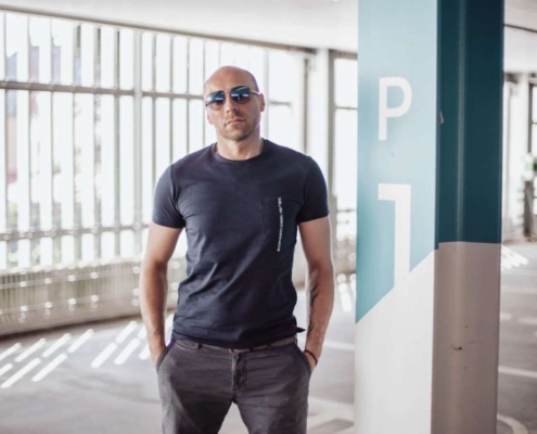 Selbstbewusst steht Callboy Carlo neben einer Säule mit der Aufschrift P1 in einem hellen, modernen Parkhaus und macht dabei eine mühelos coole Figur. Er trägt eine dunkle Sonnenbrille und ein schwarzes T-Shirt, das seinen lässigen Stil unterstreicht. Die Hände lässig in die Taschen seiner schmalen Jeans gesteckt, lehnt er mit einer entspannten, aber aufmerksamen Haltung an der Säule. Durch die großen Fenster strömt Sonnenlicht in klaren Linien und beleuchtet den polierten Betonboden und die eleganten Autos, die in dem weitläufigen Raum geparkt sind. Die Atmosphäre ist minimalistisch und modern - kaltes, weißes Licht, das von metallischen Oberflächen reflektiert wird - und bildet die perfekte urbane Kulisse für Carlos unaufdringliche Ausstrahlung.