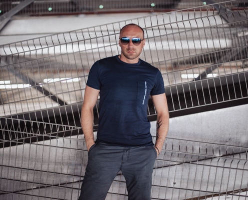 Ein Mann steht lässig vor einer Fläche aus industriellen Metallträgern und -gittern, seine Haltung ist entspannt, die Hände sind in den Taschen seiner grauen Hose versenkt. Er trägt ein marineblaues T-Shirt, das sich deutlich von dem strukturierten, metallischen Hintergrund abhebt. Eine dunkle Sonnenbrille verdeckt seine Augen und verstärkt eine Aura von lässigem Auftreten und kühlem Selbstbewusstsein, die an Callboy Carlo erinnert. Das Zusammenspiel der harten Linien des Zauns und der umgebenden Stahlkonstruktionen lässt ihn als eine Figur erscheinen, die sich in der düsteren städtischen Umgebung wohlfühlt und sich doch von ihr abhebt.