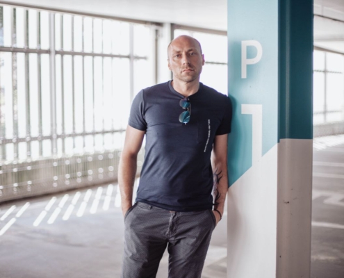 Ein Mann steht lässig in einem hell erleuchteten Parkhaus. Er hat einen rasierten Kopf und trägt ein marineblaues T-Shirt mit einer vom Kragen herabhängenden Sonnenbrille und eine graue Hose, die Hände in die Taschen gesteckt. Entspannt lehnt er an einer großen Säule mit der Aufschrift "P1". Die Szene ist minimalistisch und doch eindeutig und erinnert an den entspannten, mühelosen Stil von Callboy Carlo: kühles Selbstvertrauen durch einfache Kleidung und entspannte Haltung inmitten der Betongeometrie des Parkhauses.