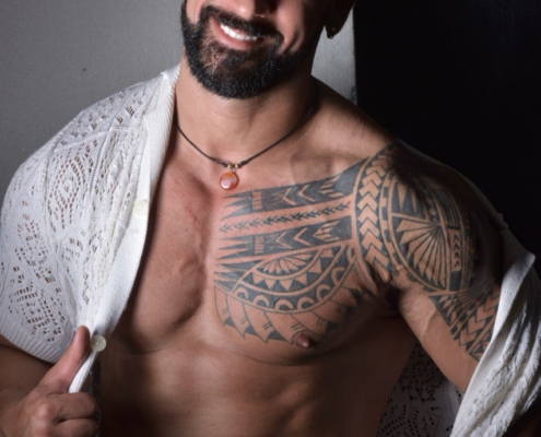 Ein lächelnder, bärtiger Mann mit dunklem Haar und einem auffälligen Tribal-Tattoo auf der Brust posiert als Callboy Amador ohne Hemd in einer offenen weißen Strickjacke.