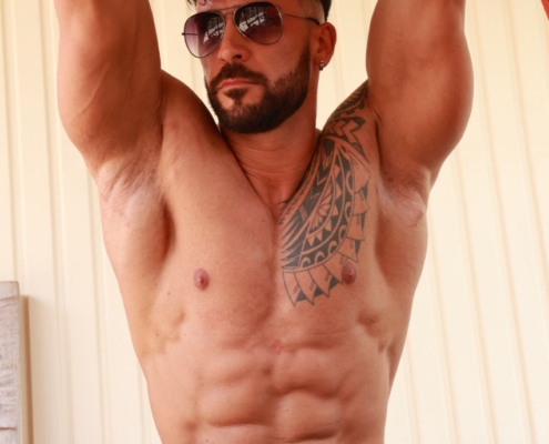 Callboy Amador, muskulös und tätowiert, mit Sonnenbrille und schwarzer Hose, hängt an einer Bar im Freien und posiert selbstbewusst an einer hellen Wand.
