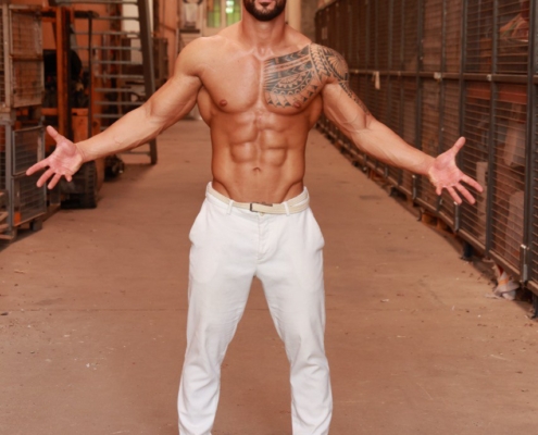 Callboy Amador, hemdsärmelig und mit sichtbaren Tattoos, steht selbstbewusst in einer Industriehalle, die Arme weit ausgebreitet, in weißen Hosen und Turnschuhen.
