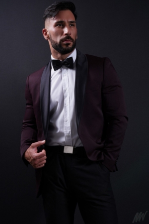 Callboy Amador, dunkelhaarig und bärtig, mit weißem Hemd, schwarzer Fliege und Satin-Lapel-Anzug, steht selbstbewusst vor einem dunklen Hintergrund.