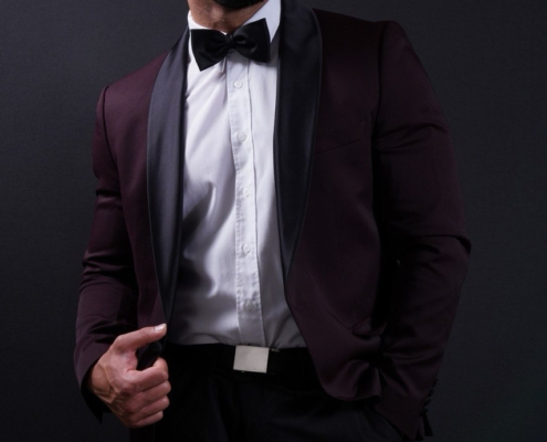 Callboy Amador, dunkelhaarig und bärtig, mit weißem Hemd, schwarzer Fliege und Satin-Lapel-Anzug, steht selbstbewusst vor einem dunklen Hintergrund.