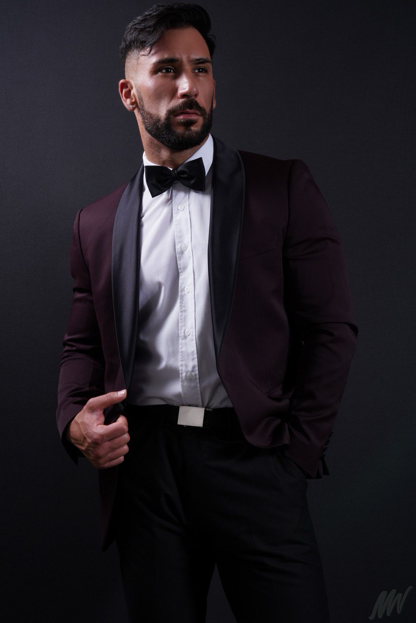 Callboy Amador, dunkelhaarig und bärtig, mit weißem Hemd, schwarzer Fliege und Satin-Lapel-Anzug, steht selbstbewusst vor einem dunklen Hintergrund.