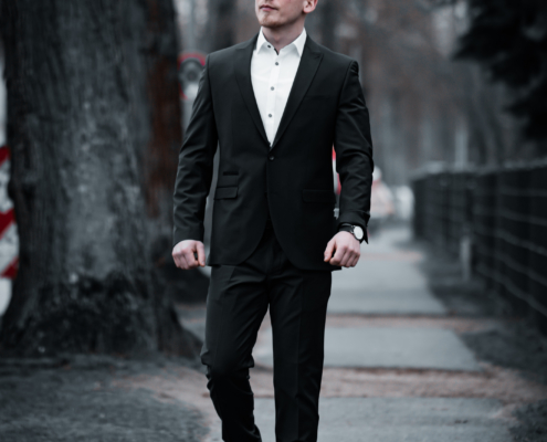 Gabriel L., tadellos gekleidet in einem eleganten schwarzen Anzug mit stilvollen braunen Schuhen, schreitet selbstbewusst über einen sonnendurchfluteten, baumgesäumten Gehweg. Sein konzentrierter Gesichtsausdruck zieht die Aufmerksamkeit auf sich und hebt sich klar vom weichen, unscharfen Hintergrund ab – ein visueller Effekt, der ihn ins Rampenlicht rückt und seine imposante Präsenz inmitten einer ansonsten ruhigen Stadtszene unterstreicht.