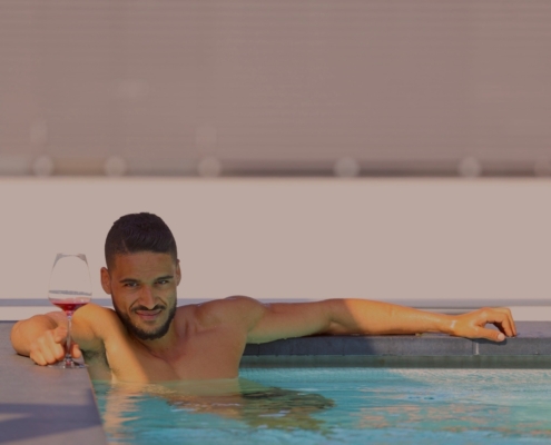 Callboy Steve liegt in der Abenddämmerung in einem ruhigen Swimmingpool. Seine durchtrainierte Figur lehnt sich an die glatten Kacheln, ein Arm ist lässig über den Beckenrand gelegt, der andere hält elegant ein Glas tiefroten Wein. Das kühle blaue Wasser plätschert sanft um ihn herum und reflektiert die goldenen Lichter der Stadt über ihm. Hinter ihm erheben sich sanft und unscharf die Türme der Stadt - das Licht schimmert durch die Panoramafenster und umrahmt diesen Moment luxuriöser Entspannung mit kosmopolitischem Charme. Steves Gesichtsausdruck ist zufrieden und ruhig; er genießt sowohl den Wein als auch die heitere Flucht inmitten des Großstadttrubels.