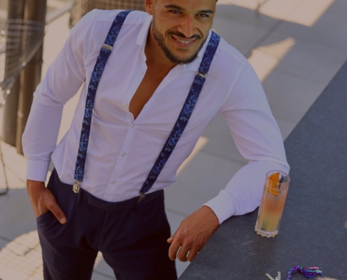 Ganz genau! Hier ist eine anschauliche Beschreibung, die auf Ihrem Szenario basiert: Callboy Steve, gekleidet in ein knackiges weißes Hemd mit auffälligen blauen Hosenträgern, steht lässig vor einem modernen, schlichten Tresen im Freien. Sein lässiges Lächeln versprüht Charme, während die Nachmittagssonne sanft auf seinem gepflegten Haar glitzert. Vor ihm auf dem Tresen steht ein lebendiger Cocktail, der mit seiner frischen Garnierung und den Kondensationsperlen Entspannung und Muße suggeriert. Neben dem Glas liegen eine modische Sonnenbrille und ein glänzender Autoschlüssel an einem auffälligen Schlüsselbund - alles subtile Hinweise auf Steves stilvolle Persönlichkeit und seinen wohlhabenden Lebensstil. Die Umgebung unter freiem Himmel verleiht dieser Momentaufnahme von selbstbewusster Kultiviertheit eine einladende Atmosphäre. Wenn Sie eine noch genauere Beschreibung oder einen anderen Verwendungszweck benötigen (z. B. eine visuelle Anregung für Kunst oder Fotografie), lassen Sie es mich wissen!.