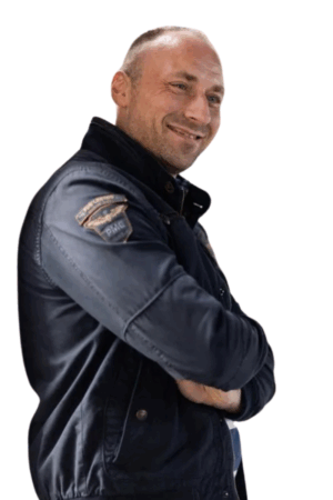 Ein Mann mit kurzen, gepflegten Haaren steht selbstbewusst vor einem schlichten weißen Hintergrund. Er trägt eine dunkle Jacke mit einem markanten Patch am Oberarm – vielleicht erinnert er an Arbeitskleidung oder eine Fliegerjacke – und hat die Arme lässig vor der Brust verschränkt. Seine Haltung ist entspannt und dennoch selbstbewusst. Er lächelt herzlich, den Blick zur Seite gerichtet, als würde er einen Insiderwitz erzählen oder etwas Lustiges aus dem Off beobachten. Sein Gesichtsausdruck und seine Haltung strahlen den selbstbewussten Charme aus, der mit Callboy Carlo assoziiert wird.