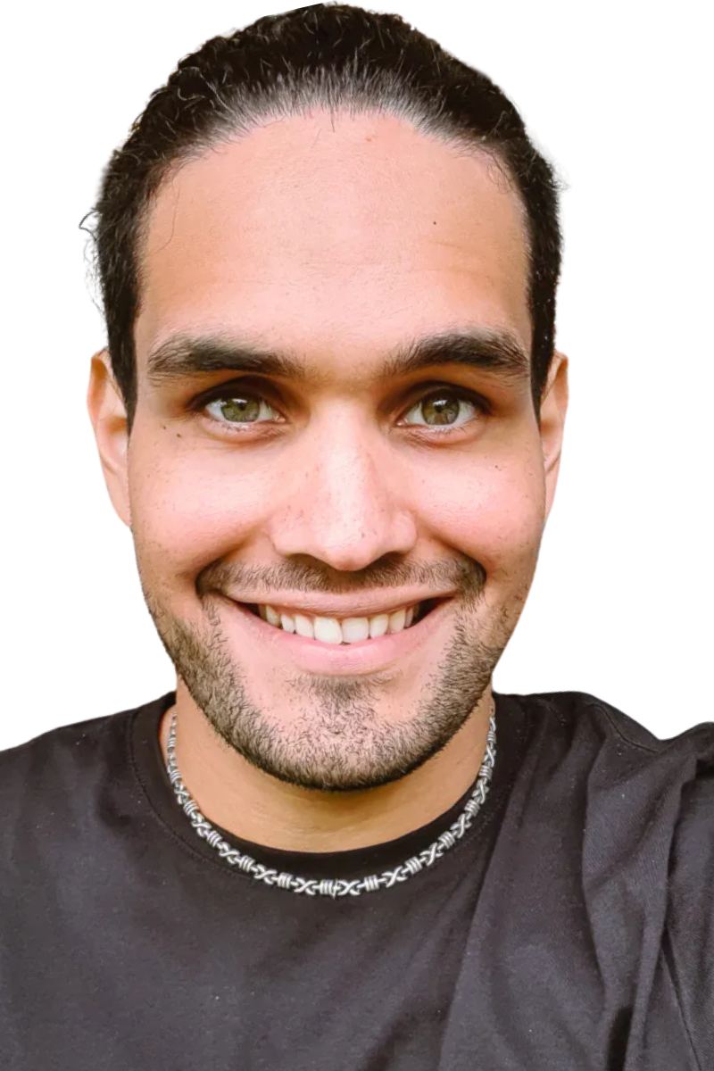 Ein junger Mann mit zurückgekämmtem dunklem Haar, hellen Augen und gestutztem Bart lächelt in die Kamera. Er trägt ein schwarzes Hemd und eine Perlenkette. Der Hintergrund ist schlicht und weiß und unterstreicht seinen freundlichen Gesichtsausdruck. Sein Look ist lässig und dennoch gepflegt, wobei der schlichte Schmuck seinem Erscheinungsbild Persönlichkeit verleiht.