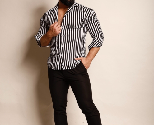Ein Mann mit dunklem Haar und Bart steht selbstbewusst vor einem schlichten Hintergrund und strahlt das Charisma von Callboy Kaan aus. Er trägt ein auffälliges schwarz-weiß gestreiftes Hemd, kombiniert mit einer engen schwarzen Hose und eleganten schwarzen Schuhen. Seine Hände ruhen lässig in den Taschen, oder er rückt seine Manschetten leicht zurecht und strahlt Selbstbewusstsein aus. Der Gesamtlook ist modern, mutig und raffiniert – perfekt für Callboy Kaans charakteristischen Stil.